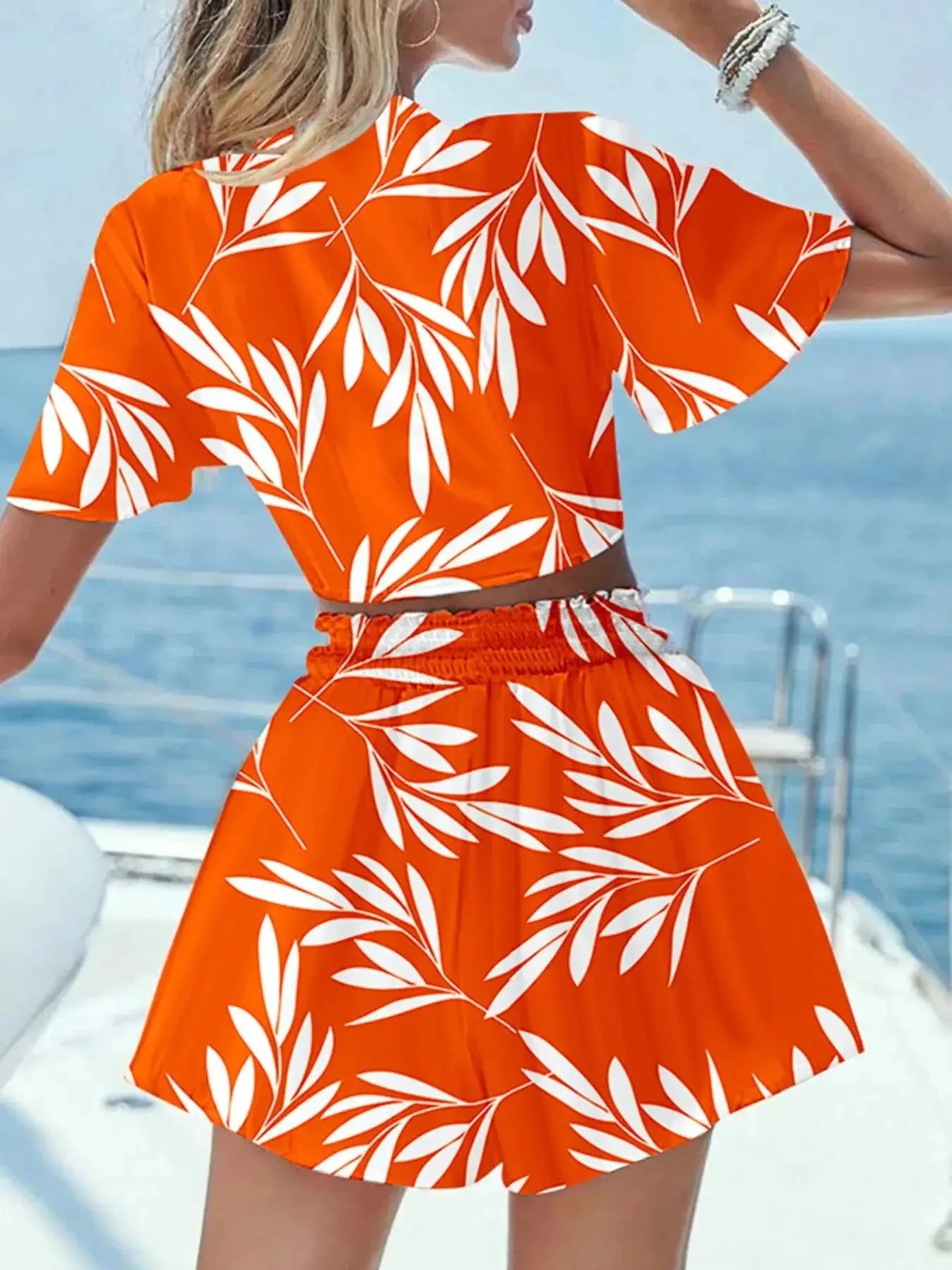Model wearing an orange printed tie front top and shorts set with white leaf design, on a boat.