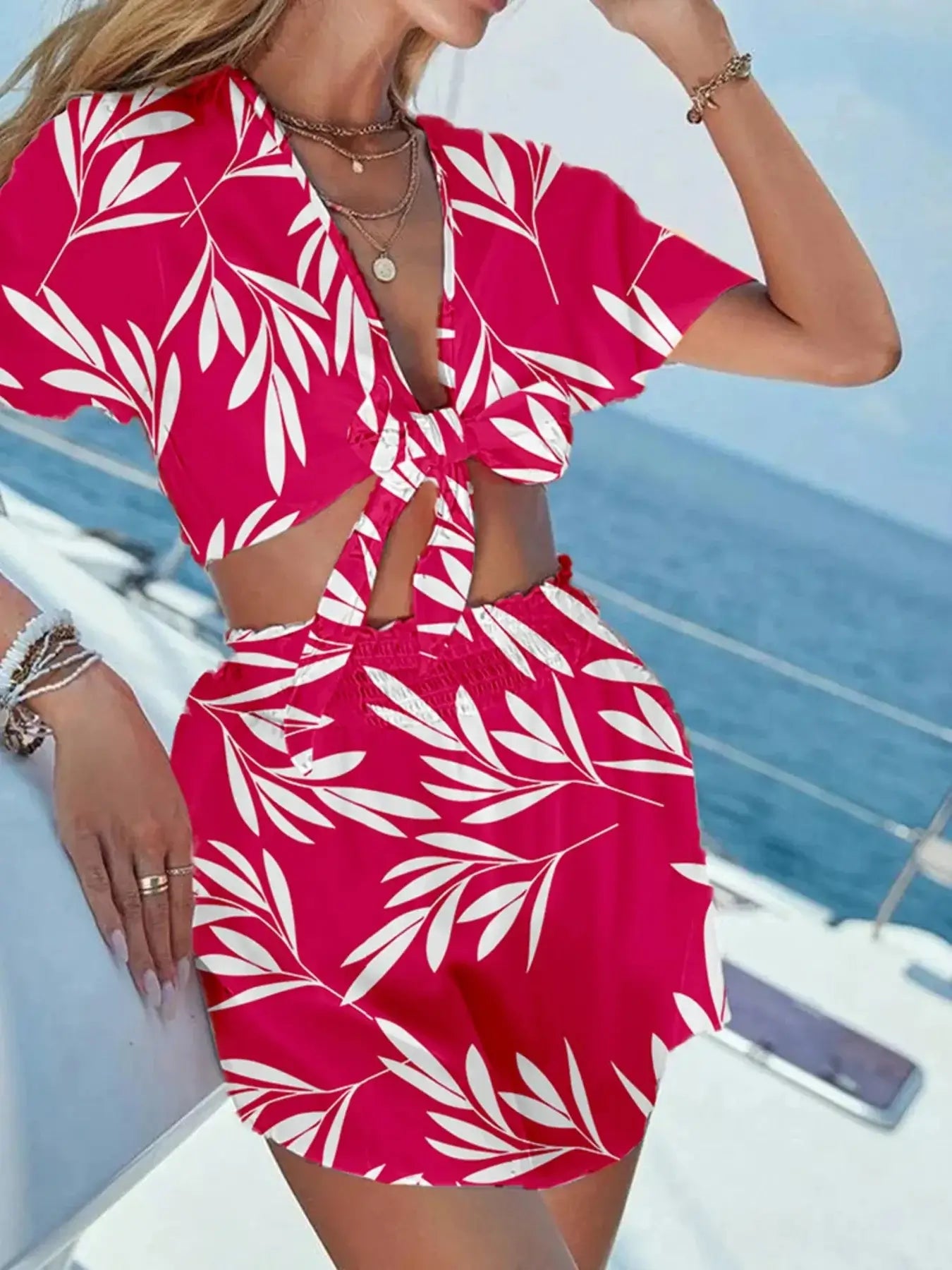 Model wearing a printed tie front top and shorts set in bright pink with white leaf patterns, posed by the seaside.